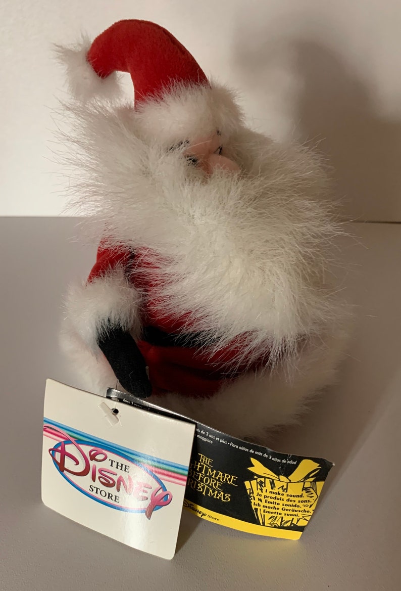 May include: A plush Santa Claus figure with a red hat and suit, white faux fur trim, and black boots. The figure has a tag from The Disney Store, with text that reads "The Nightmare Before Christmas".