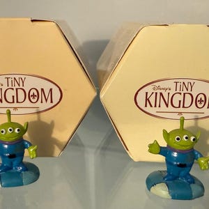 May include: Two Disney's Tiny Kingdom collectible figures of the Little Green Men from Toy Story. Each figure is green with a blue uniform and is standing on a blue and white base. The figures are in front of a light yellow box with the Tiny Kingdom logo.