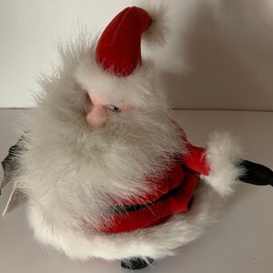 May include: A plush Santa Claus figure, featuring a red hat and suit trimmed with white faux fur. The figure has a white beard and a pink face. Black boots complete the festive look. A tag is attached to the figure.