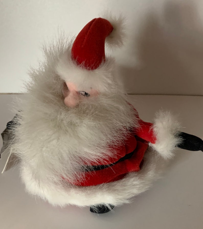 May include: A plush Santa Claus figure, featuring a red hat and suit trimmed with white faux fur. The figure has a white beard and a pink face. Black boots complete the festive look. A tag is attached to the figure.
