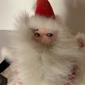 May include: A plush Santa Claus doll with a red hat and a long white beard. The doll has a pink face and black eyes. The beard is made of fluffy white material. The doll is a festive decoration.