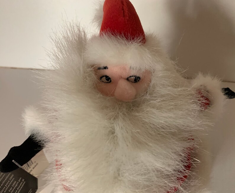 May include: A plush Santa Claus doll with a red hat and a long white beard. The doll has a pink face and black eyes. The beard is made of fluffy white material. The doll is a festive decoration.
