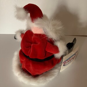 May include: A plush Santa Claus figure, viewed from the side, wearing a red velvet suit with white faux fur trim. The figure has a black belt and a tag from The Disney Store.