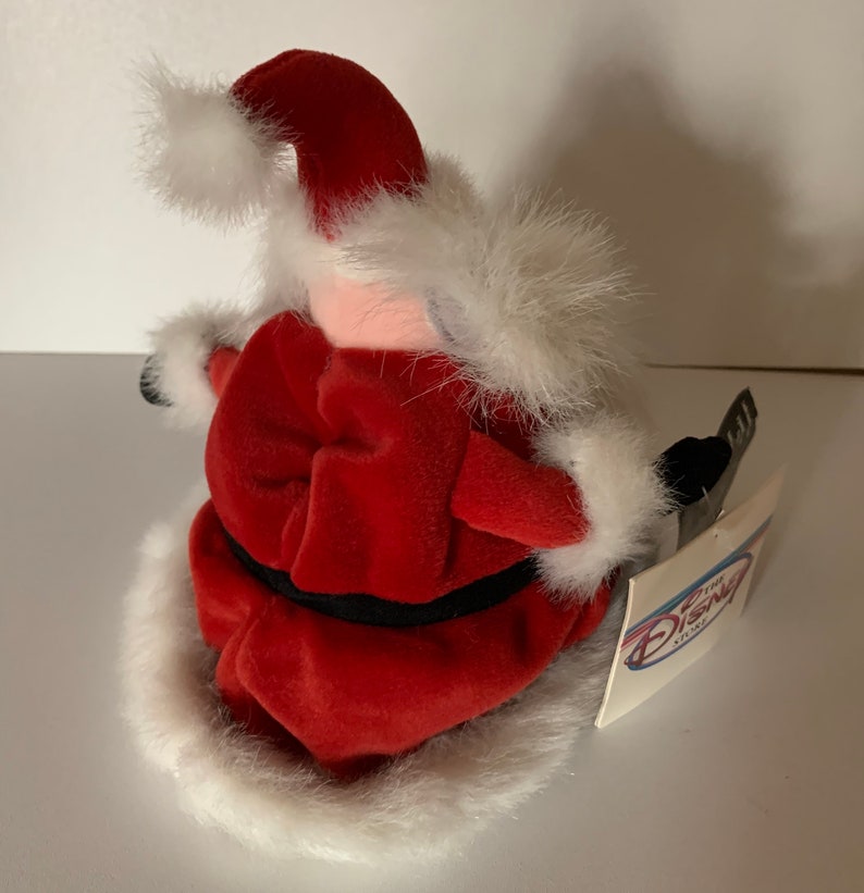 May include: A plush Santa Claus figure, viewed from the side, wearing a red velvet suit with white faux fur trim. The figure has a black belt and a tag from The Disney Store.