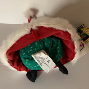 May include: A plush toy of a character from "The Nightmare Before Christmas" wearing a red Santa Claus outfit with white faux fur trim. The toy has black feet and a green body with a holly pattern. A tag is attached.