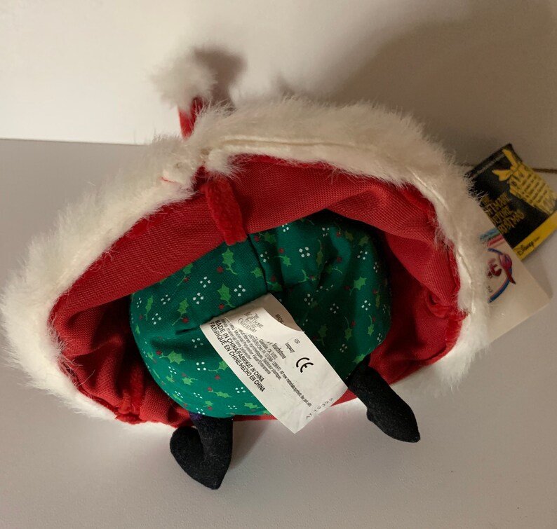 May include: A plush toy of a character from "The Nightmare Before Christmas" wearing a red Santa Claus outfit with white faux fur trim. The toy has black feet and a green body with a holly pattern. A tag is attached.
