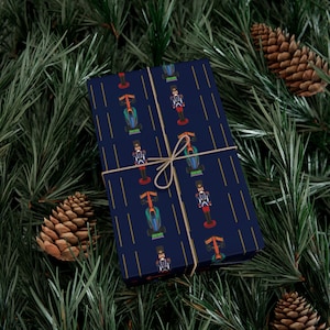 May include: Dark blue wrapping paper with a repeating pattern of race cars and nutcrackers, tied with twine. The paper is decorated with vertical gold lines, and is surrounded by pine needles and pine cones.