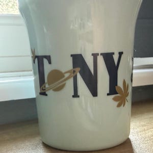Op de afbeelding: Een witte keramische mok met de zwarte tekst TONY, waarbij de letter O is vervangen door een gouden planeetgrafiek en een gouden bladgrafiek rechts van de letter Y is geplaatst. De mok heeft een glad, cilindervormig lichaam met een licht uitlopende rand.