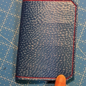 May include: A blue leather notebook with red stitching. The notebook is closed and lying flat on a blue and white gridded surface.