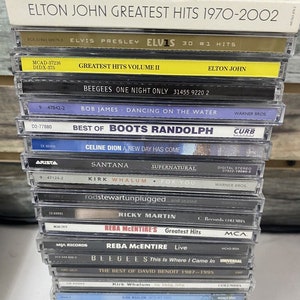 May include: A stack of 20 compact discs (CDs) featuring various artists and albums. The CDs are arranged vertically, with the titles and artist names visible. Some of the artists include Elton John, Elvis Presley, The Eagles, and Celine Dion.
