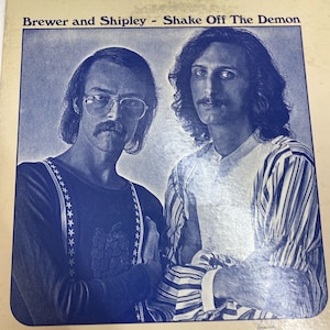 May include: A vintage record album cover featuring a blue and white photo of two men. The album title is "Brewer and Shipley - Shake Off The Demon".