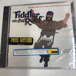 May include: A CD case with a black background and a colorful image of a man in a blue hat and brown jacket dancing. The title "Fiddler on the Roof" is written in large, white letters. The text "ORIGINAL MOTION PICTURE SOUNDTRACK RECORDING" is written in smaller white letters above the title. The text "SH/FIDDLER ON/SOUNDTRAC 725754//F113006//216" is written in white letters on a yellow tag.
