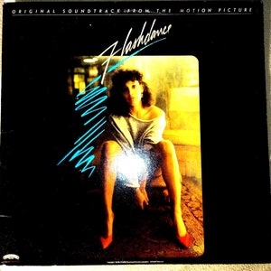 May include: A black vinyl record album cover with the title "Flashdance" in white text with a blue outline. The cover features a photo of a woman sitting on a couch with a yellow background. The woman is wearing a gray sweater and red shoes.