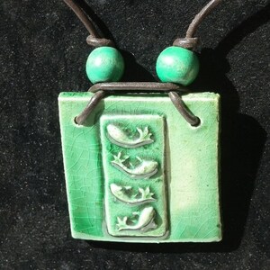 Può includere: Un ciondolo in ceramica verde con un design di quattro pesci stilizzati, appeso a un cordino di cuoio marrone con perline verdi.