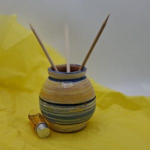 K&ouml;nnte beinhalten: Eine Keramik-Diffuser-Vase mit einem gelben, blauen und wei&szlig;en Streifenmuster. Die Vase hat drei Holzst&auml;bchen im Inneren. Eine kleine Glasflasche mit goldener Kappe steht vor der Vase.