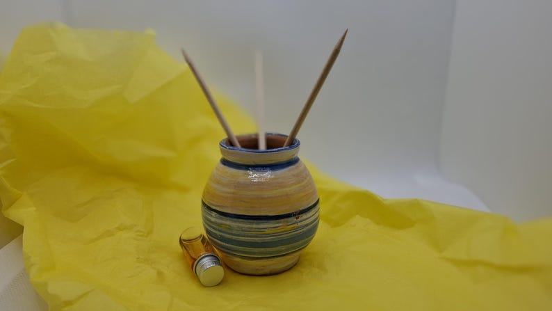 K&ouml;nnte beinhalten: Eine Keramik-Diffuser-Vase mit einem gelben, blauen und wei&szlig;en Streifenmuster. Die Vase hat drei Holzst&auml;bchen im Inneren. Eine kleine Glasflasche mit goldener Kappe steht vor der Vase.