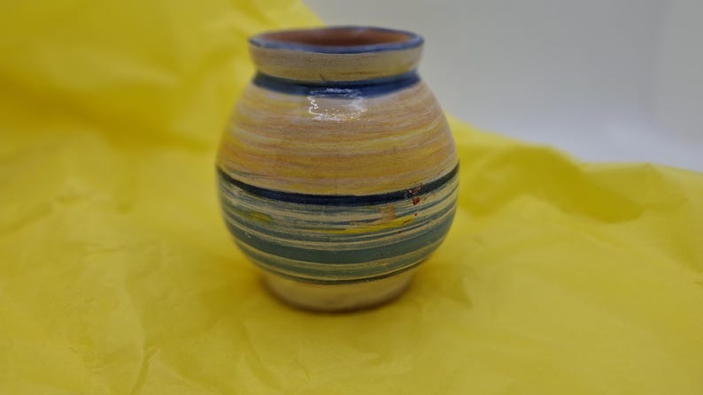 K&ouml;nnte beinhalten: Eine kleine, handbemalte Keramikvase mit rundem K&ouml;rper und einer weiten &Ouml;ffnung. Die Vase ist mit blauen, gelben und wei&szlig;en Streifen verziert.
