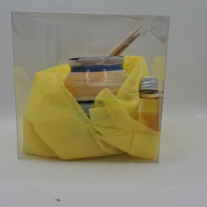 K&ouml;nnte beinhalten: Eine transparente Plastikbox mit einem kleinen Keramikgef&auml;&szlig; mit blauen und gelben Streifen, das in gelbes Seidenpapier eingewickelt ist. Eine kleine Glasflasche mit silbernem Verschluss und klarer Fl&uuml;ssigkeit ist ebenfalls enthalten.