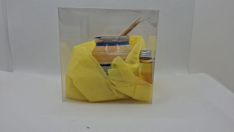 K&ouml;nnte beinhalten: Eine transparente Plastikbox mit einem kleinen Keramikgef&auml;&szlig; mit blauen und gelben Streifen, das in gelbes Seidenpapier eingewickelt ist. Eine kleine Glasflasche mit silbernem Verschluss und klarer Fl&uuml;ssigkeit ist ebenfalls enthalten.