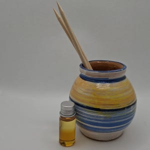 Może przedstawiać: Ceramiczny dyfuzor z żółto-niebiesko-białym wzorem w paski. Dyfuzor ma dwa drewniane patyczki w środku i małą butelkę olejku eterycznego obok.