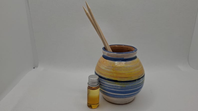 K&ouml;nnte beinhalten: Ein Keramikdiffusor mit einem gelben, blauen und wei&szlig;en Streifenmuster. Der Diffusor hat zwei Holzst&auml;bchen im Inneren und eine kleine Flasche &auml;therisches &Ouml;l daneben.