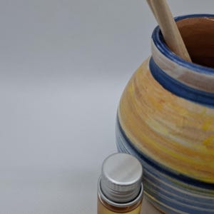 K&ouml;nnte beinhalten: Eine blaue und gelbe gestreifte Keramikvase mit einem Holzr&uuml;hrer im Inneren. Eine kleine silberne Flasche mit einer klaren Fl&uuml;ssigkeit befindet sich im Vordergrund.