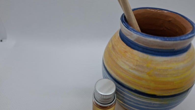K&ouml;nnte beinhalten: Eine blaue und gelbe gestreifte Keramikvase mit einem Holzr&uuml;hrer im Inneren. Eine kleine silberne Flasche mit einer klaren Fl&uuml;ssigkeit befindet sich im Vordergrund.