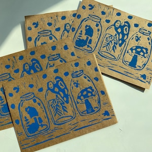 Op de afbeelding: Vier bruine kaarten met blauwe inkt-illustraties van potten met daarin paddenstoelen, bladeren en een kleine boom. De kaarten hebben een herhalend patroon van kleine blauwe stippen.