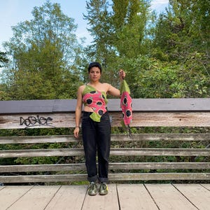 May include: A person wearing a black top with a pink and green patterned design, black pants, and green shoes. The person is standing on a wooden bridge and holding a pink and green patterned scarf.