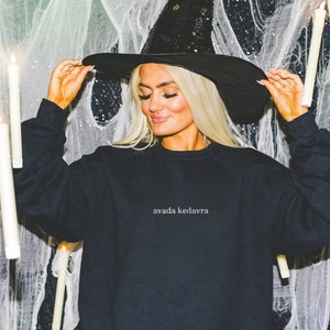Peut inclure: Une femme portant un chapeau de sorcière noir et un sweat-shirt noir avec le texte "avada kedavra" en blanc. Elle se tient devant un fond blanc avec des bougies blanches.