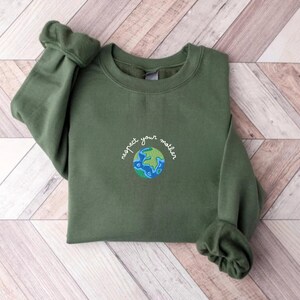 Puede incluir: Una sudadera verde con el texto "respect your mother" bordado en la parte delantera, con una imagen estilizada de la Tierra debajo del texto.