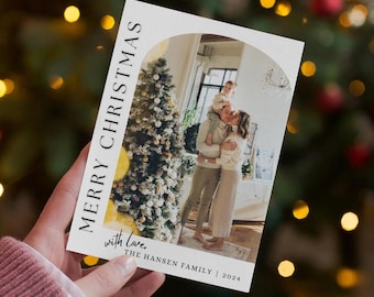 Pode incluir: Um cartão de Natal branco com uma foto de uma família em pé em frente a uma árvore de Natal decorada. O texto no cartão diz "Feliz Natal com amor, A família Hansen | 2024".
