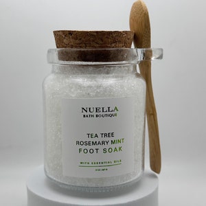 Puede incluir: Un tarro de vidrio transparente lleno de cristales blancos para baño de pies, coronado con una tapa de corcho. Una pequeña cuchara de madera descansa a un lado. La etiqueta dice "NUELLA BATH BOUTIQUE TEA TREE ROSEMARY MINT FOOT SOAK WITH ESSENTIAL OILS".