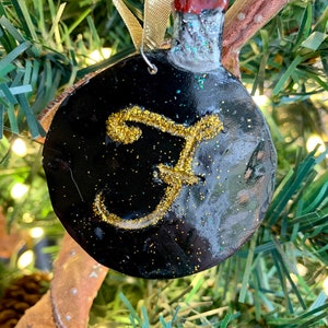 May include: Black and gold Christmas ornament shaped like a bottle with a gold letter "F" on the front. The ornament is hanging on a Christmas tree.