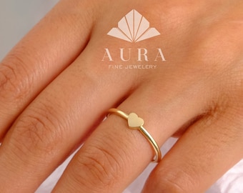May include: A simple gold ring with a small heart-shaped design. The ring is worn on a finger with a light pink nail polish.