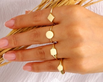 May include: A hand wearing four gold rings. The rings are different shapes, including a square, a circle, and two rectangles. The rings are stacked on the fingers.