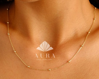 May include: A delicate gold chain necklace with small round beads. The necklace is worn by a person with a light complexion. The brand name "AURA FINE JEWELLERY" is visible in the image.