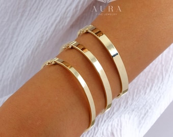 May include: Four gold bangle bracelets stacked on a person's arm. The bracelets are all different sizes and have a smooth, polished finish. The brand name "AURA FINE JEWELRY" is visible on the image.