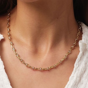 May include: A gold-toned necklace with a repeating pattern of linked segments. The necklace has a two-tone design, with gold and silver-colored elements. The jewelry is displayed against a white background.