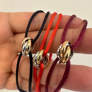 Könnte beinhalten: Drei Armbänder mit ineinandergreifenden Ringen in Silber-, Gold- und Roségoldtönen. Jedes Armband besteht aus einer dünnen, glatten Kordel in Schwarz, leuchtendem Orange oder Dunkelburgund, die durch die Mitte der drei Metallringe gefädelt ist.