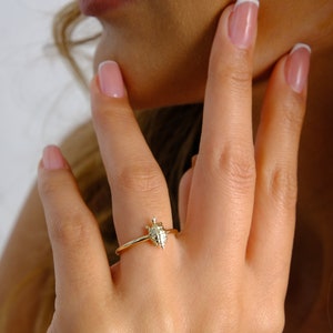 Puede incluir: Un anillo de oro con un pequeño diseño de tortuga. El anillo está en el dedo de una persona.