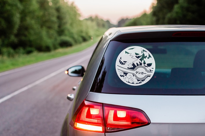 May include: A white car with a black and white sticker on the back window. The sticker is a circle with a Japanese character in the center. The car is parked on a road with trees in the background.