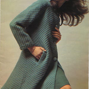 May include: A teal-colored, textured coat with large buttons and a knee-length design. The coat features a unique pattern and a pocket. The model is wearing a bracelet and a ring.