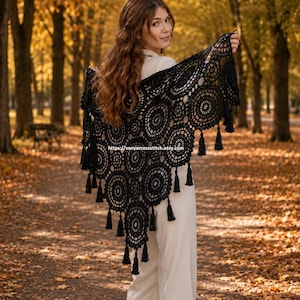 May include: A black crocheted shawl with a circular design and tassels. The shawl is worn over the shoulders. The background shows a park with trees and fallen leaves.