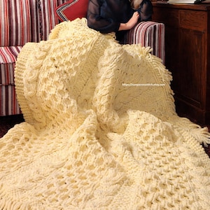 Könnte beinhalten: Eine cremefarbene, handgestrickte Decke mit strukturiertem Muster und Fransen. Die Decke ist über ein Sofa mit rot-weißem Streifenmuster drapiert. Das Design der Decke umfasst Zopfmuster und Wabenmuster.