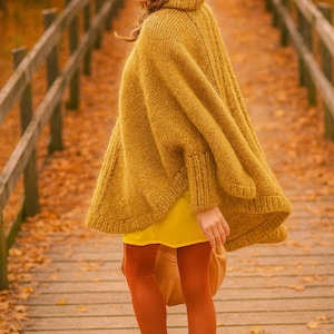 Könnte beinhalten: Eine Frau trägt einen senfgelben Strickponcho, ein gelbes Kleid und rostfarbene Strumpfhosen. Sie trägt braune Stiefel mit Absatz und steht auf einer Holzbrücke mit Herbstlaub.