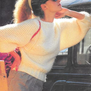 May include: A woman wearing a cream-colored, knitted sweater with a crew neck and long, puffy sleeves. The sweater has a ribbed hem and cuffs. The image also shows the word "Beehive" in white text at the top. The woman is wearing sunglasses and gray pants.