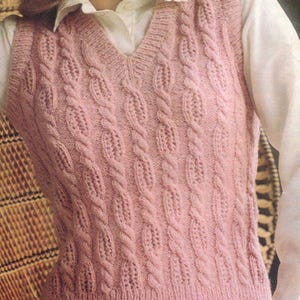 Puede incluir: Un chaleco de punto rosa claro con diseño de trenzas y cuello en V. El chaleco presenta un patrón de trenzas detallado que recorre verticalmente la parte delantera. La prenda está hecha de un hilo suave y texturizado, y se usa sobre una camisa blanca.