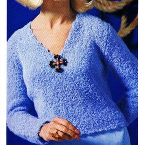 May include: A woman wearing a light blue knit sweater with a V-neck and a matching skirt. The sweater has a decorative brooch. The image is from a vintage fashion advertisement. The word "COMMODORE" is printed at the bottom.
