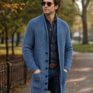 Könnte beinhalten: Ein langer, blauer Strickcardigan mit Knopfleiste und zwei Vordertaschen. Der Cardigan wird über einem dunkelblauen Hemd und Jeans getragen. Die Person trägt eine Sonnenbrille und einen braunen Gürtel.